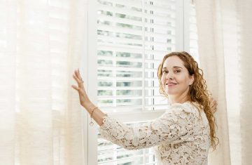 Kobieta przy oknie z białymi shuttersami i jasnymi zasłonami